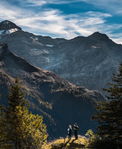 Two people stand on a grassy hilltop surrounded by trees, with large, rocky mountains and a partly cloudy sky in the background.