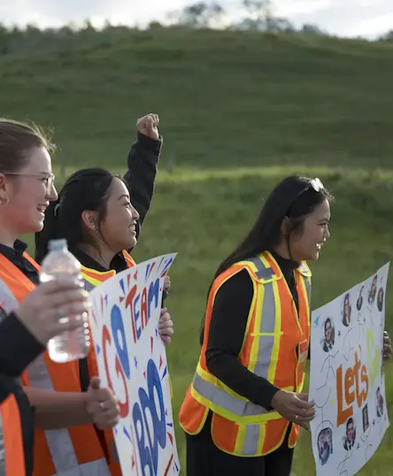 cheering-on-kananaskis-relay-race-k100-mile-relay-resized Three people in reflective vests stand outdoors, holding colorful signs and a water bottle. One person raises a fist. The background is a grassy hill under a cloudy sky. The scene suggests support for an event or team.