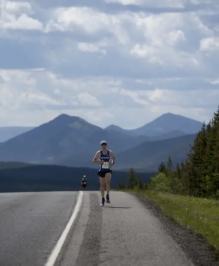 A runner in athletic gear and a race bib runs alone on a rural road with mountains, trees, and a partly cloudy sky in the background. Another person is visible farther down the road.