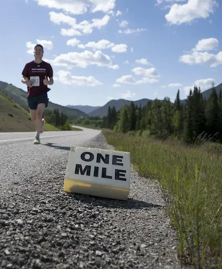 one-mile-marker-k100-mile-relay-resized A person running on a paved road in a rural, mountainous area passes a sign on the ground that reads “ONE MILE” with trees and blue sky in the background.
